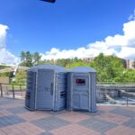 A gray ADA-compliant portable toilet next to a gray handicap-accessible portable toilet on a city walkway