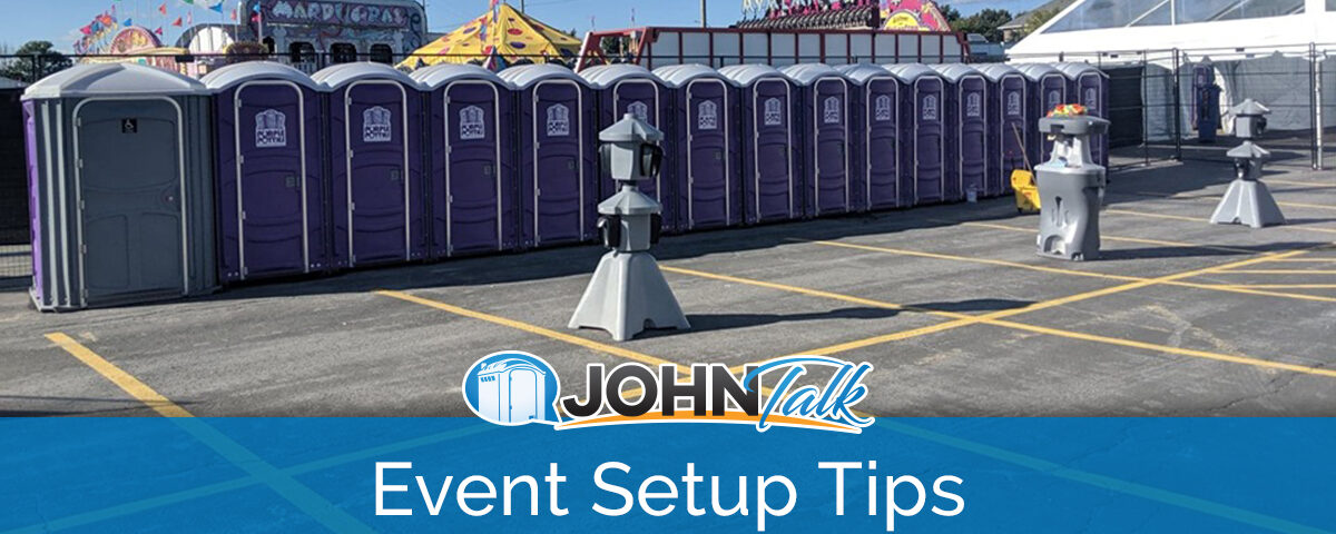 A long row of purple portable toilets, a sink and two hand sanitizing stations with a carnival in the background