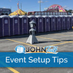 A long row of purple portable toilets, a sink and two hand sanitizing stations with a carnival in the background